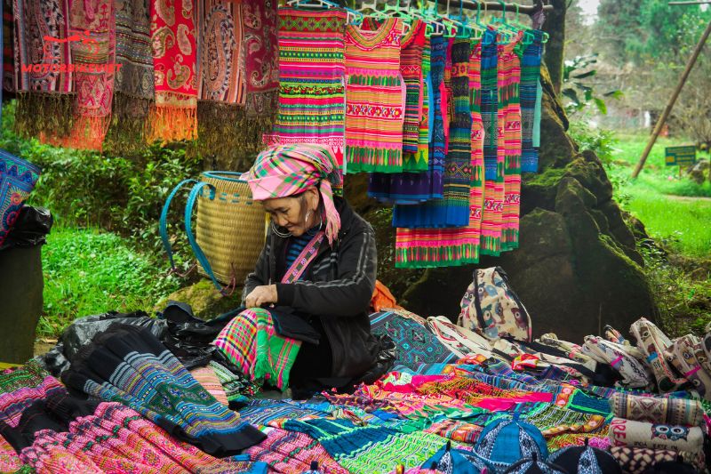 A woman in traditional attire sewing amidst colorful embroidered textiles at an outdoor market