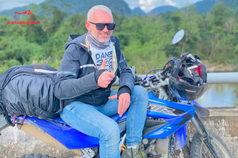 Man giving a thumbs up while sitting on a blue motorbike in a mountain landscape